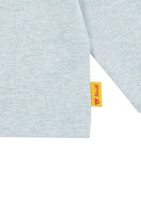 Hellblaue Baumwollbluse mit langen Ärmeln und sichtbarem gelben Etikett mit dem Markennamen. Strukturierter Stoff und angenähte Saumdetails.