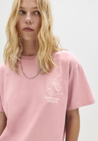 Ung kvinde med blonde bølger, iført en pink t-shirt med et granatæblemotiv og teksten "SANGUINE TOUCH", ser let mod venstre.