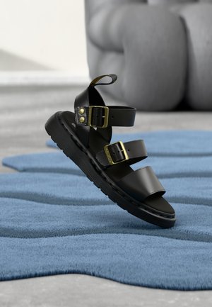 Schwarze Ledersandale mit drei Riemen und goldenen Schnallen, schwebend über einer blau strukturierten Oberfläche dargestellt.