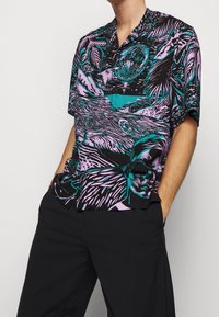 Chemise à manches courtes présentant un imprimé tropical vibrant en turquoise et violet sur un fond noir, avec une coupe décontractée et un col.