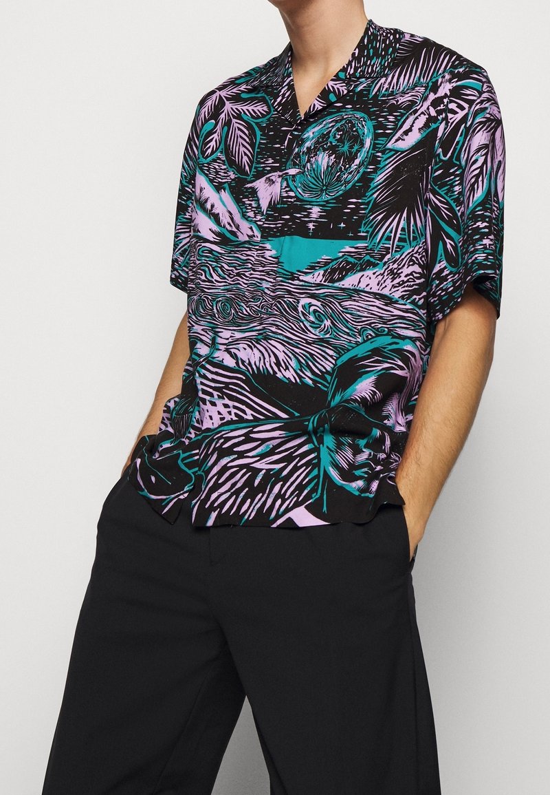 Chemise à manches courtes présentant un imprimé tropical vibrant en turquoise et violet sur un fond noir, avec une coupe décontractée et un col.