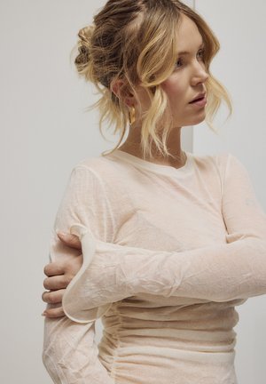 Femme aux cheveux blonds coiffés en chignon, portant un haut beige transparent à manches longues, tenant son bras gauche avec sa main droite, regardant vers la droite.