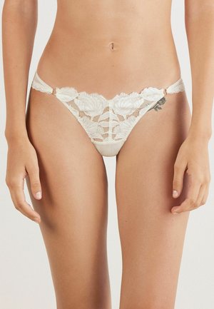 Vrouw in witte bloemenkant bikini-achtige onderbroek met dunne zijkanten en een zichtbare heup tattoo aan de rechterkant.