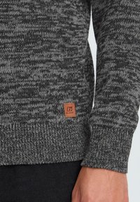 Gros plan sur la manche et le torse d'un pull en tricot gris avec un petit patch en cuir marron de la marque "Blend" près de l'ourlet, porté avec un pantalon noir.