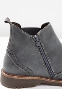 Šedá kožená kotníková bota s bočním zipem, elastickou vsadkou a dekorativním prošíváním. Má strukturovaný povrch a hnědou podrážku.