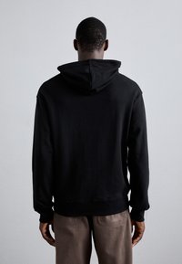 Hoodie noir avec une poche kangourou, capuche à cordon et poignets côtelés. Porté avec un pantalon marron, montrant un tissu doux et texturé.