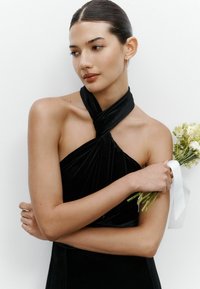 Rochie din catifea neagră cu design asimetric și pliere. Subiectul ține un buchet împachetat în panglică, pe un fundal alb simplu.