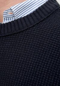 Suéter de punto azul marino con un patrón texturizado, que presenta un cuello acanalado y una camisa a rayas azul claro y blanca asomando por debajo.