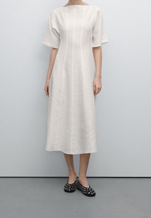 Femme portant une robe midi blanche avec des manches volantées et des chaussures noires à motifs circulaires blancs, se tenant devant un fond uni.