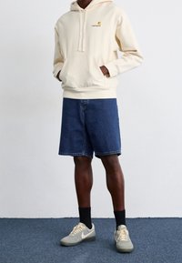 Felpa crema con tasca frontale e logo, abbinata a pantaloni corti in denim blu, calze nere e sneakers grigie con dettagli beige.