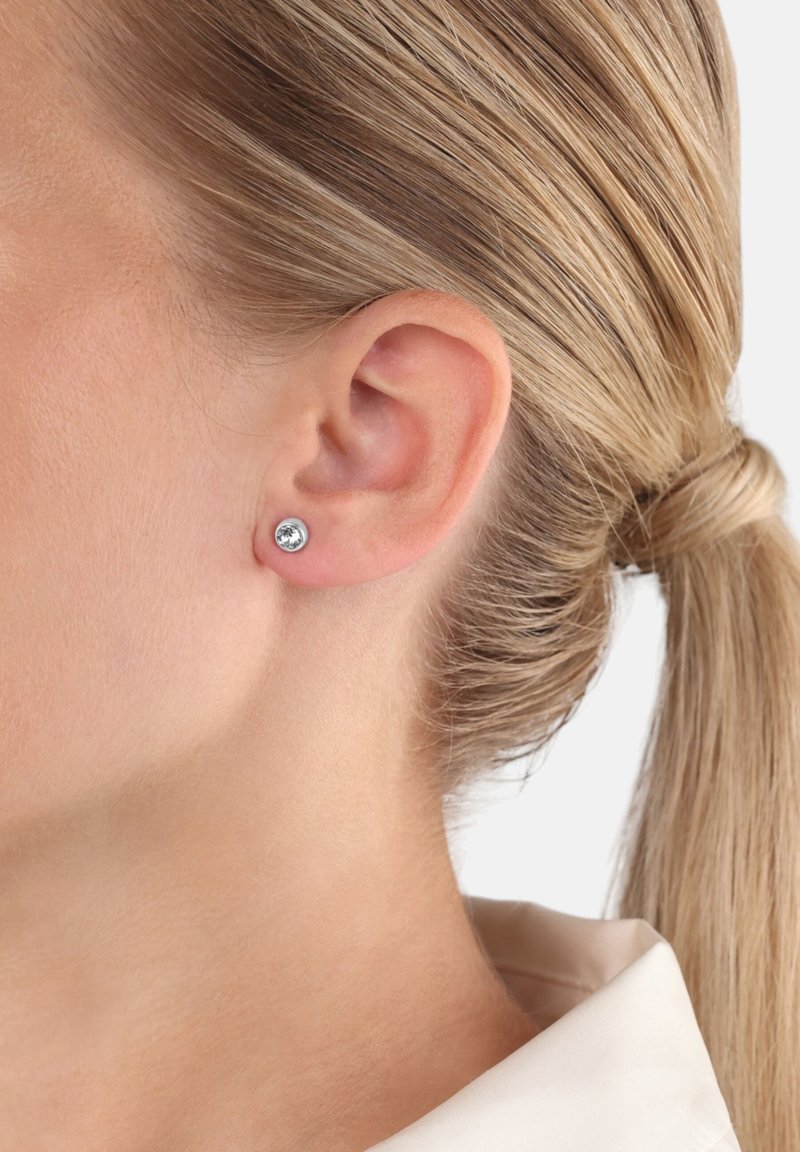 Silverörhänge med en klar ädelsten, rund design och polerad yta, bärs på en modells öra och visar enkelhet och elegans.