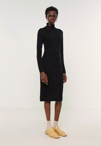 Μαύρο πλεκτό φόρεμα με εφαρμοστή σιλουέτα, μακριά μανίκια και ψηλό λαιμό. Λείος υφασμάτινος σχεδιασμός και μήκος μέχρι τη γάμπα. Φοριέται με μπεζ κάλτσες και κίτρινα παπούτσια.