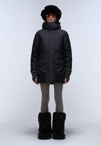 Svart puffjacka med hög krage och två framfickor, matchad med grå leggings och höga svarta fodrade stövlar. Solglasögon med bruna linser.
