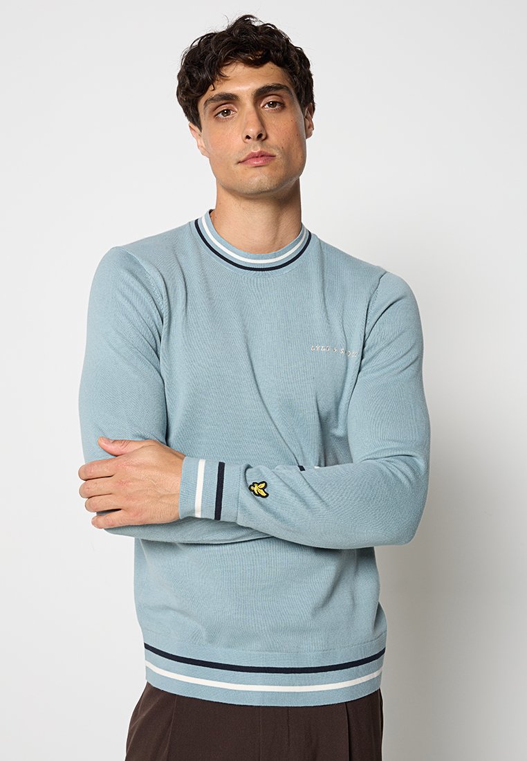 Maglione blu a coste con colletto e polsini a righe in contrasto. Presenta un logo giallo ricamato sulla manica sinistra. Design aderente.