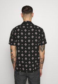 Korte mouwen zwarte shirt met een geometrisch patroon in wit en rood, voorzien van een kraag en een ontspannen pasvorm. Glad stoffen textuur.