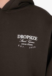 Gros plan sur un hoodie marron foncé avec un texte blanc : "DROPSIZE Street Game DREAM CHASERS EST. 2020" sur la poitrine.