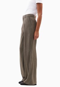Kalhoty s širokými nohavicemi z texturované hnědé látky, s vysokým pasem, předními sklady a bočními kapsami, v kombinaci s černými botami.