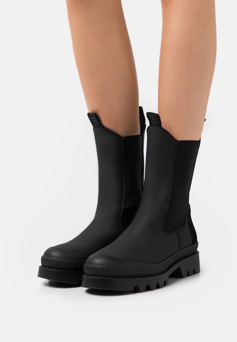 Bottes en caoutchouc noires mi-mollet avec semelles épaisses et rainurées portées sur des jambes nues sur un fond blanc uni.