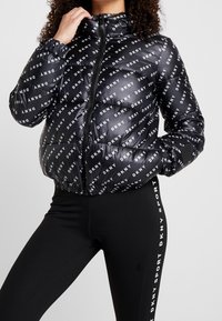 Černá péřová bunda s opakujícím se bílým logem "DKNY", texturovaná tkanina, zipový uzávěr, vysoký límec a elastické manžety; doplněná černými legínami.