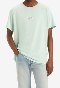 Mint green short-sleeve T-shirt made of soft cotton, featuring a black Levi's logo patch on the chest and a relaxed fit. Paired with light blue jeans.