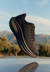 Chaussure de sport noire avec semelle épaisse et dessus en maille, flottant au-dessus d'une surface réfléchissante avec des montagnes et des arbres flous en arrière-plan.