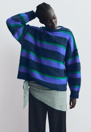 Knitted sweater in dark blue with purple and green stripes, loose fit, wide sleeves. Paired with a grey draped top and black pants.