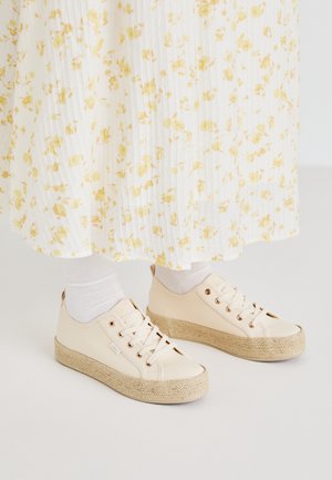Pieds portant des baskets compensées couleur crème avec semelles tissées et chaussettes blanches, associés à une jupe blanche à motifs de fleurs jaunes.
