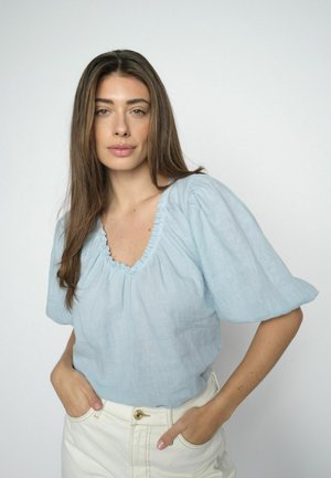 Hosszú barna hajú nő világoskék, fodros ujjú blúzban és fehér nadrágban, kezeit a zsebében tartva, egyszerű háttér előtt áll.