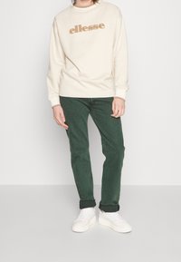 Vit sweatshirt med guld "ellesse"-logotyp, kombinerad med mörkgröna jeans och vita sneakers. Avslappnad passform och texturerade material genom hela outfiten.