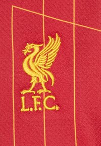 Tejido rojo con el escudo bordado en amarillo del Liverpool Football Club, que presenta un pájaro mítico y las letras "L.F.C." debajo.