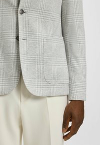 Jasnoszara marynarka w kratę z teksturowanej tkaniny, na dwa guziki, z kieszenią. Noszona z jasnymi spodniami.