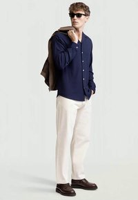 Camisa azul-marinho com texturas e botões, calças bege, sapatos castanhos e uma jaqueta castanha colocada sobre o ombro. Óculos de sol pretos completam o visual.