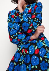 Blommig klänning i svart tyg med stora blåa och röda rosenmönster. Har långa ärmar med elastiska manschetter och en rund halsringning.