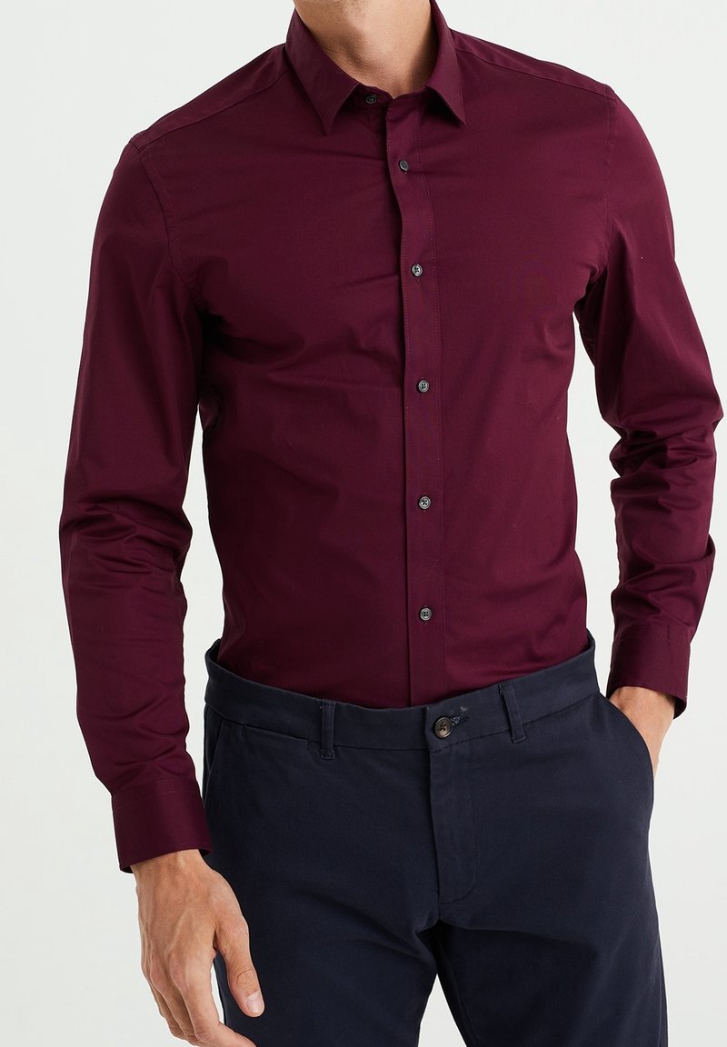 Burgundy skjort med en åtsittande design, långa ärmar, knäppkrage och mörka knappar, i kombination med marinblå byxor. Slät textur.