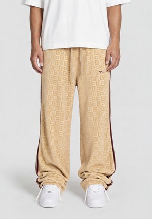 Pantaloni beige strutturati con motivo geometrico, strisce laterali scure, indossati con sneakers bianche e camicia bianca, mostrati dalla vita ai piedi.