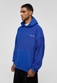 Blauwe hoodie van textuurstof, voorzien van een voortas, geribbelde manchetten en een trekkoordkap. "Eternity Project" logo in wit.