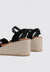 Sandalias de cuña de ante negro con un tacón de yute texturizado, que cuentan con una correa en el tobillo y una punta redondeada. La suela es plana y tiene agarre de goma.