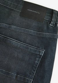 Sötét farmernadrág sima textúrával, hátsó zsebbel és egy fekete bőr címkével, amelyen a "DENIM SUPPLY Co." felirat található a derék résznél.