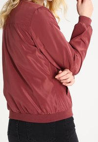 Burgundská bomber bunda s hladkým, lesklým povrchem, žebrovanými manžetami a pasem. Disponuje jednoduchým, klasickým designem a bočními kapsami.