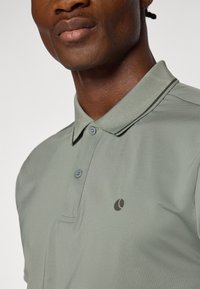 Olivengrønn polo-skjorte laget av teksturert stoff, med en placket med to knapper og en liten logo på brystet.