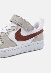 Vit sneaker med grå läderdetaljer, brun Nike swoosh, texturerad sula och kardborreband. Utrustad med vita snören och ventilerande foder.