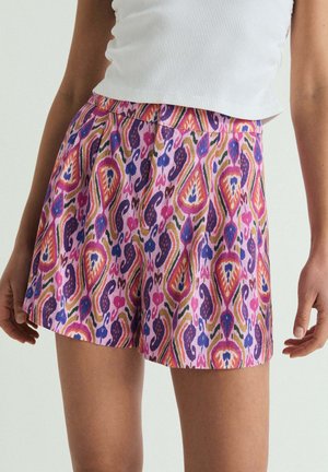 Mujer que lleva pantalones cortos estampados de colores con diseños en rosa, púrpura, naranja y azul combinados con una camiseta blanca sin mangas.