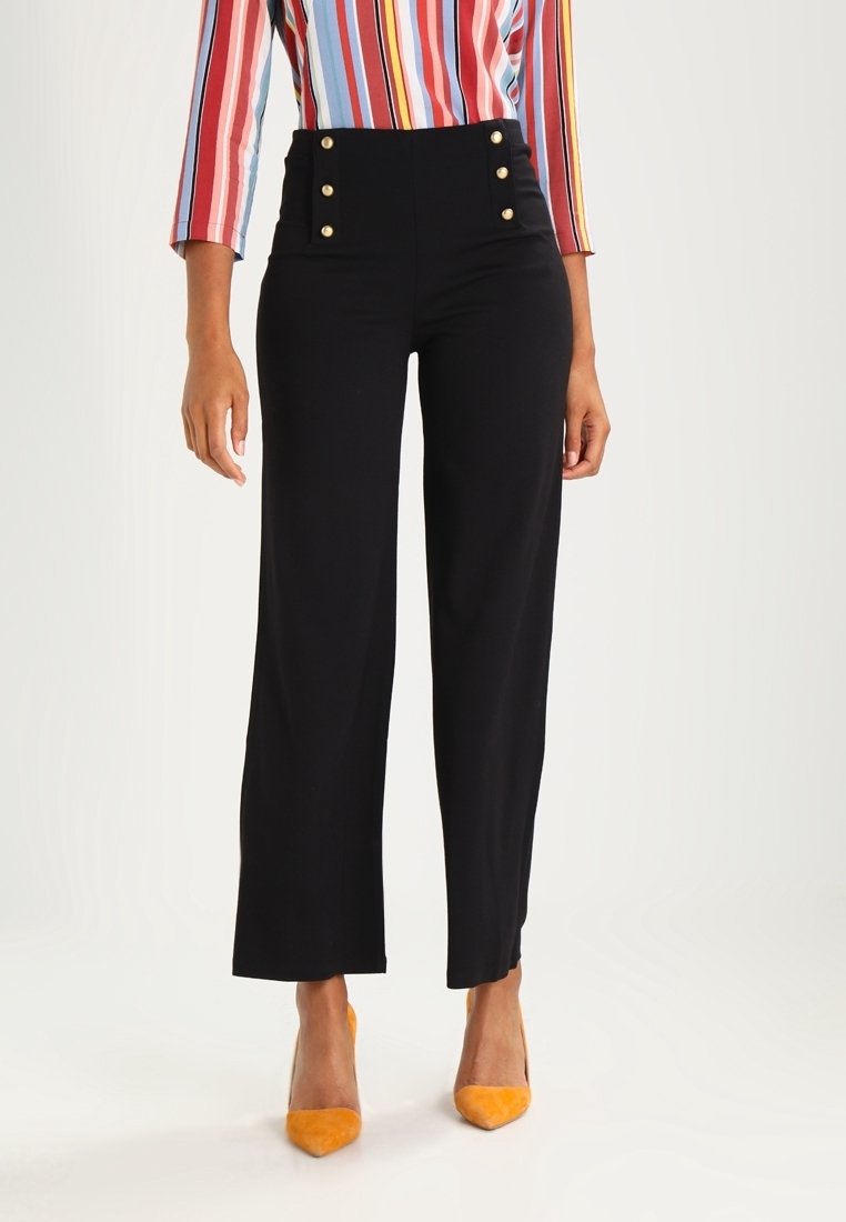 Femme portant un pantalon noir taille haute avec des boutons dorés, associé à un haut à manches longues rayé coloré et des chaussures pointues orange.