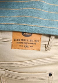 Pantalones de mezclilla color beige claro con una etiqueta de cuero que dice "Petrol Denim Driven Since 1989." El tejido presenta una textura y costuras visibles.