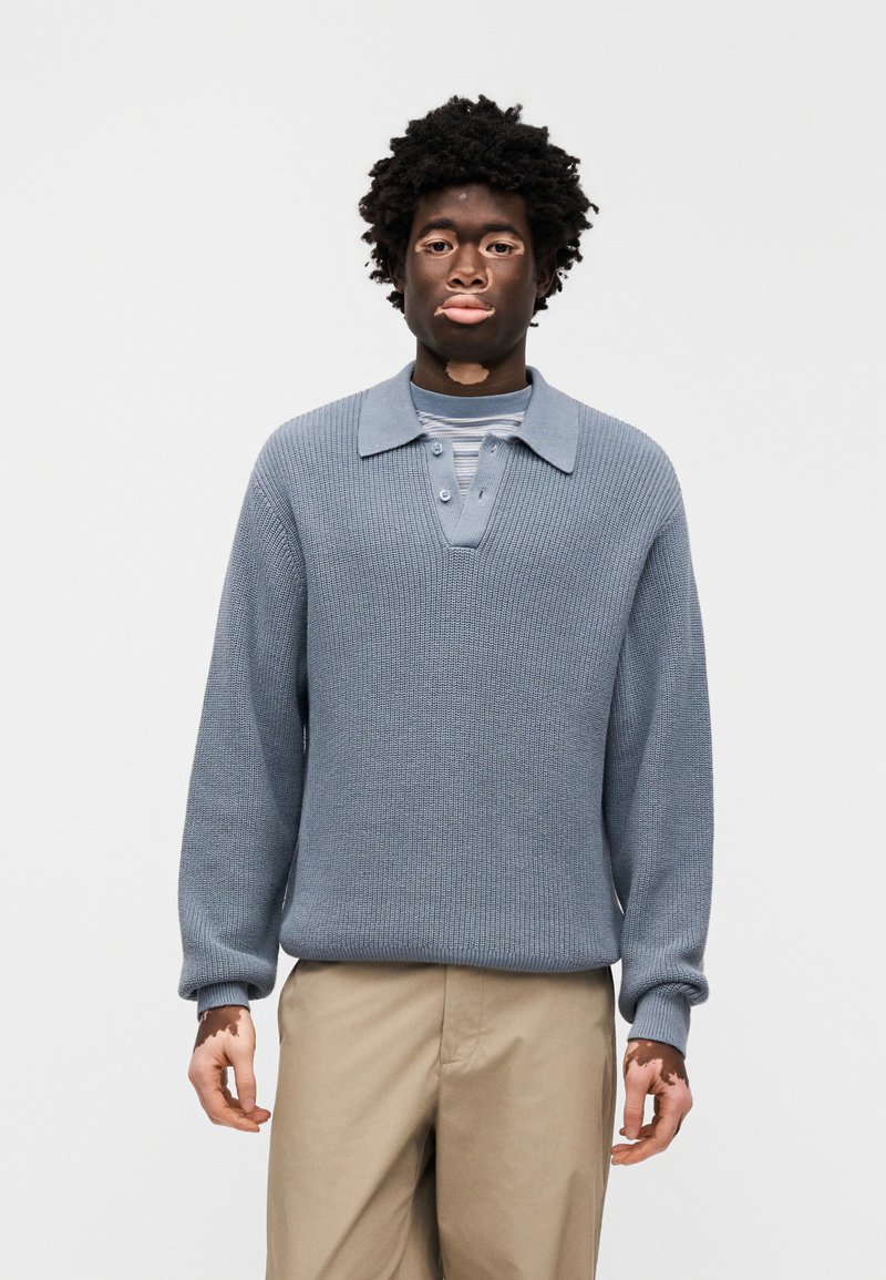 Lyseblå strikket pullover med krave, knaplukning og ribstrikkede manchetter. Båret over en langærmet, stribet skjorte.