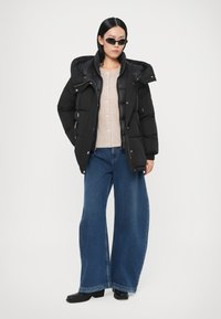 Must pufferjakk kõrge kraega, beež nööpidega kampsun, laia säärega sinised teksad ja mustad platvormsaapad. Aksessuaaride hulka kuuluvad tumedad päikeseprillid.