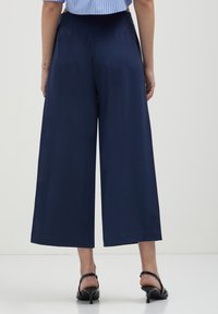 Donna che indossa pantaloni a palazzo blu navy a vita alta e corta, con sandali neri con tacco, in piedi su uno sfondo bianco.