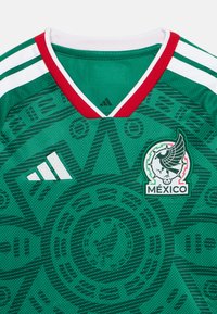 Πράσινη ποδοσφαιρική φανέλα με κόκκινο και λευκό V-λαιμό. Φέρει το λογότυπο της Adidas και το έμβλημα του Μεξικού. Υφή υφάσματος με κυκλικά σχέδια και λεπτομέρειες.