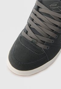 Σκούρο γκρι αθλητικό παπούτσι με σουέτ επάνω μέρος, γκρι κορδόνια, διάτρητο μπροστινό μέρος και λευκή καουτσούκ σόλα. Σημαντική λεπτομέρεια λογότυπου στο πλάι.