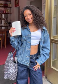 Jeansjacka med avslappnad passform, ljusblå färg och detalj med knappar; matchad med en vit crop top och mörka vida jeans. Håller en öppen bok.